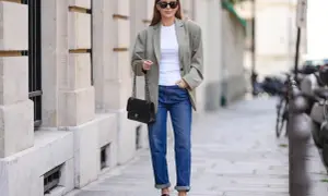 Kobieta w jeansach i oversize'owej marynarce damskiej, z czarną torebką na łańcuszku, prezentuje miejski szyk.