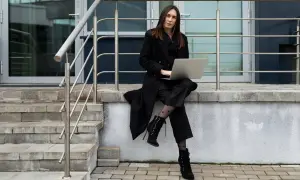 Elegancka kobieta w czarnym płaszczu, z laptopem na kolanach, prezentuje stylowe dodatki: wzorzyste rajstopy i sznurowane botki.