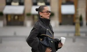 Elegancka kobieta w okularach i ciemnej kurtce puchowej, z torebką Chanel i kawą na wynos. Stylowe kurtki puchowe to podstawa jesiennych stylizacji.