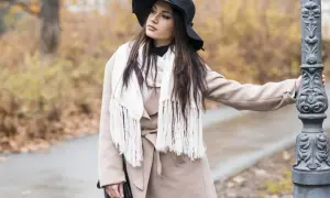 Elegancka kobieta w beżowym płaszczu i czarnym kapeluszu, z białym, frędzlowym szalem, idealnie pasującym do beżowego płaszcza, opiera się o latarnię.