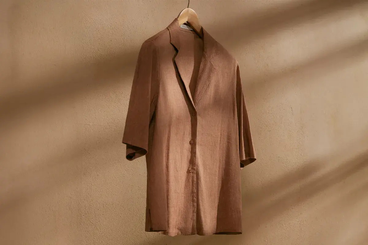 Brązowa, lniana koszula na drewnianym wieszaku. Idealna baza do letnich stylizacji, dodaje lekkości i elegancji.