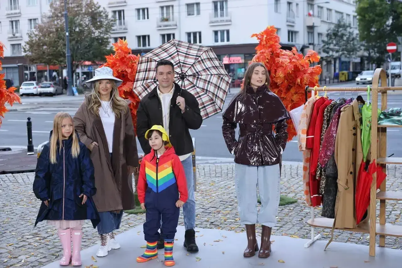 Rodzina prezentuje stylowe stylizacje na deszczowe dni. Dzieci w kolorowych kaloszach i kurtkach, dorośli w płaszczach i błyszczącej kurtce.