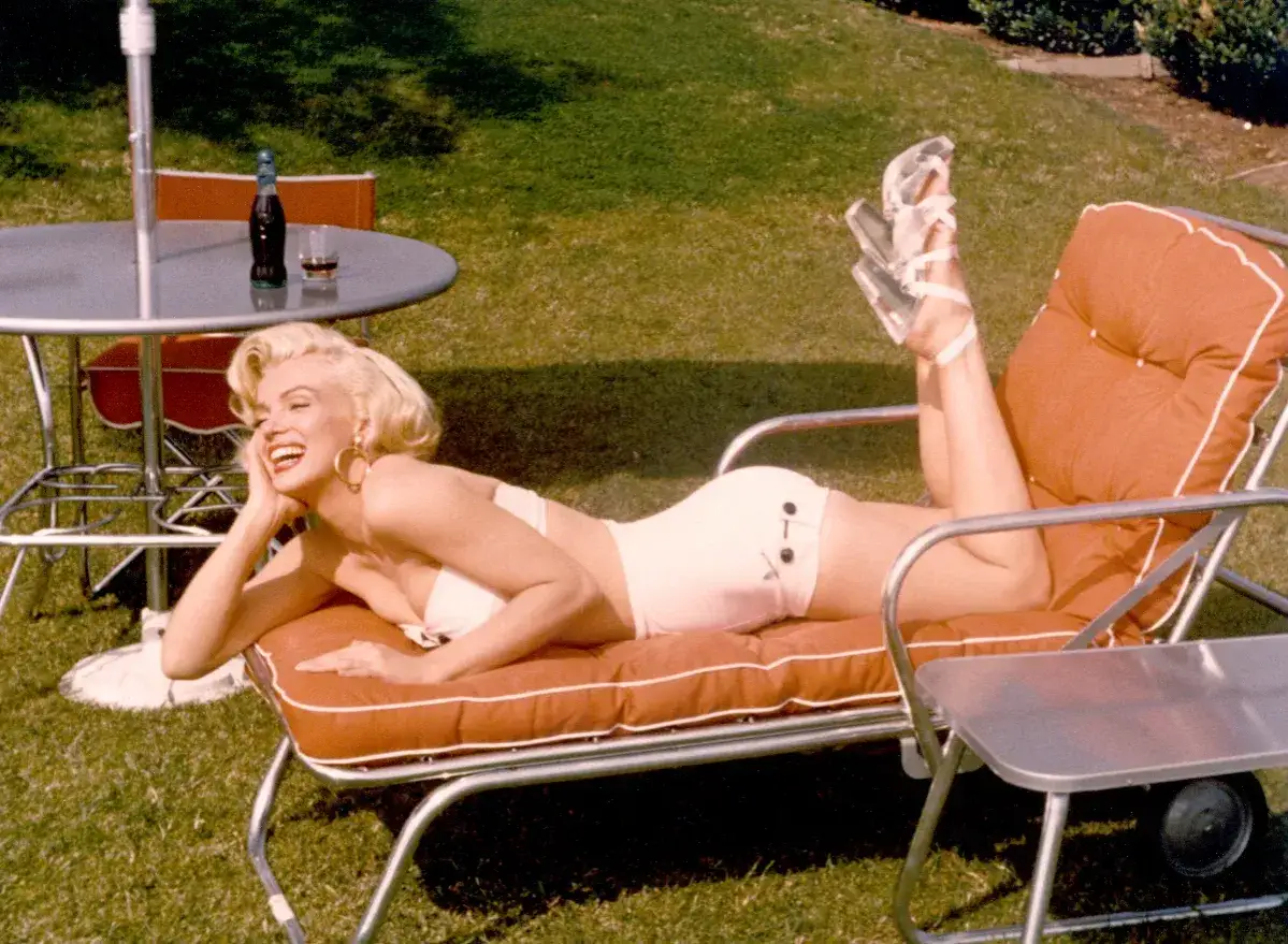 Marilyn Monroe w białym bikini, leżąca na leżaku w ogrodzie, z nogami uniesionymi w górę. Jej sylwetka jest ikoniczna.