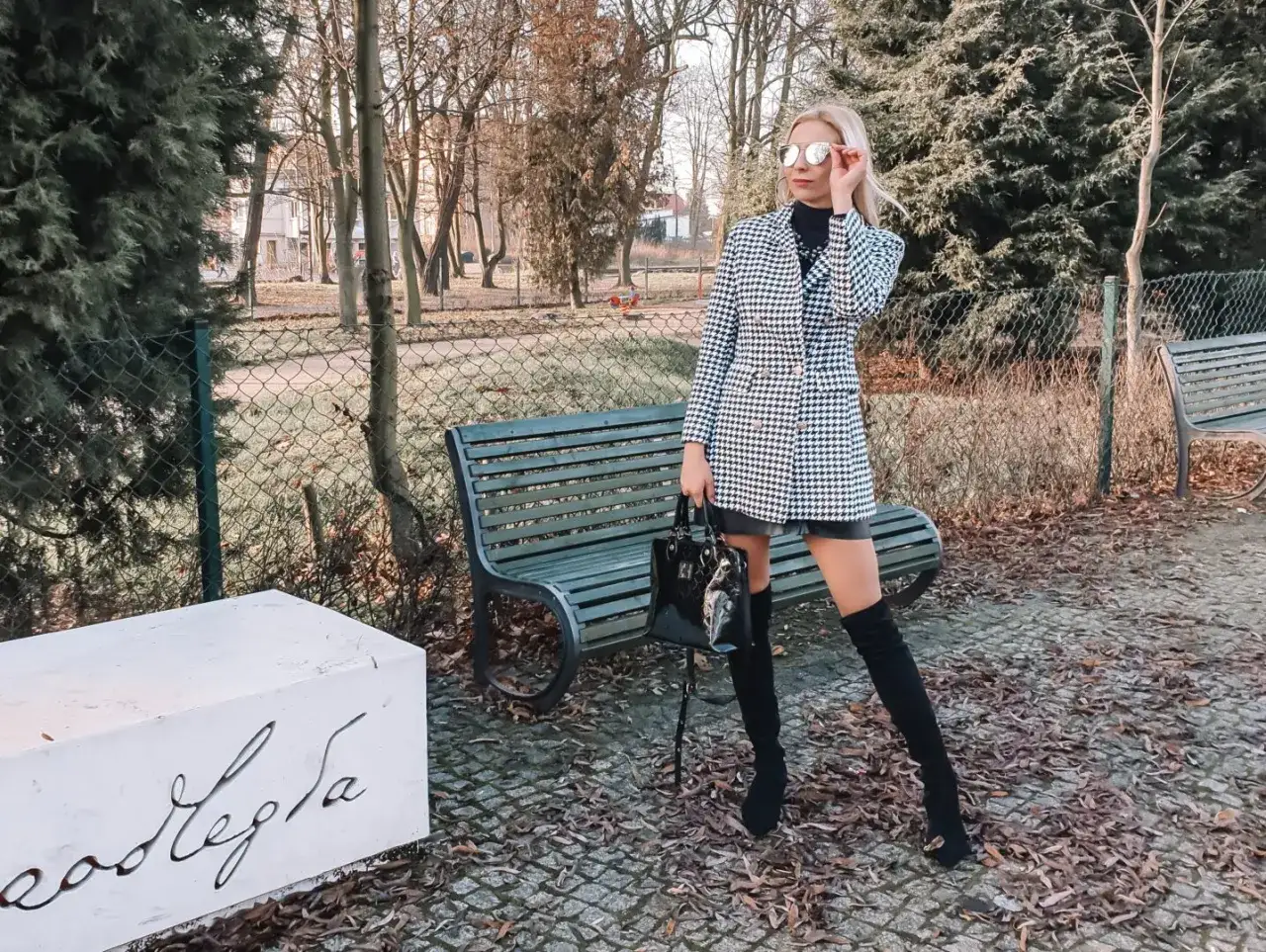 Elegancka kobieta w płaszczu w pepitkę i kozakach prezentuje modne stylizacje na tle parku.