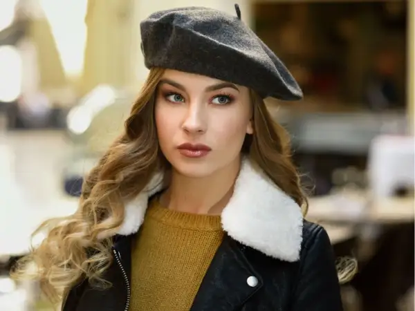 Elegancka kobieta w grafitowym beretu, z burzą blond loków i w czarnej ramonesce. Dowiedz się, jak nosić beret damski, by wyglądać stylowo.